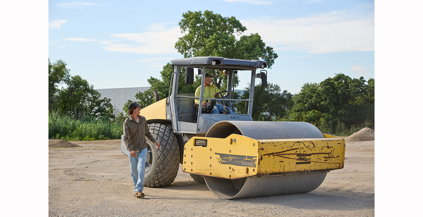 A Dreamworx employee drives a yellow Atlas Copco CA2500 soil compactor over a dirt lot, with dust rising around the large front roller.